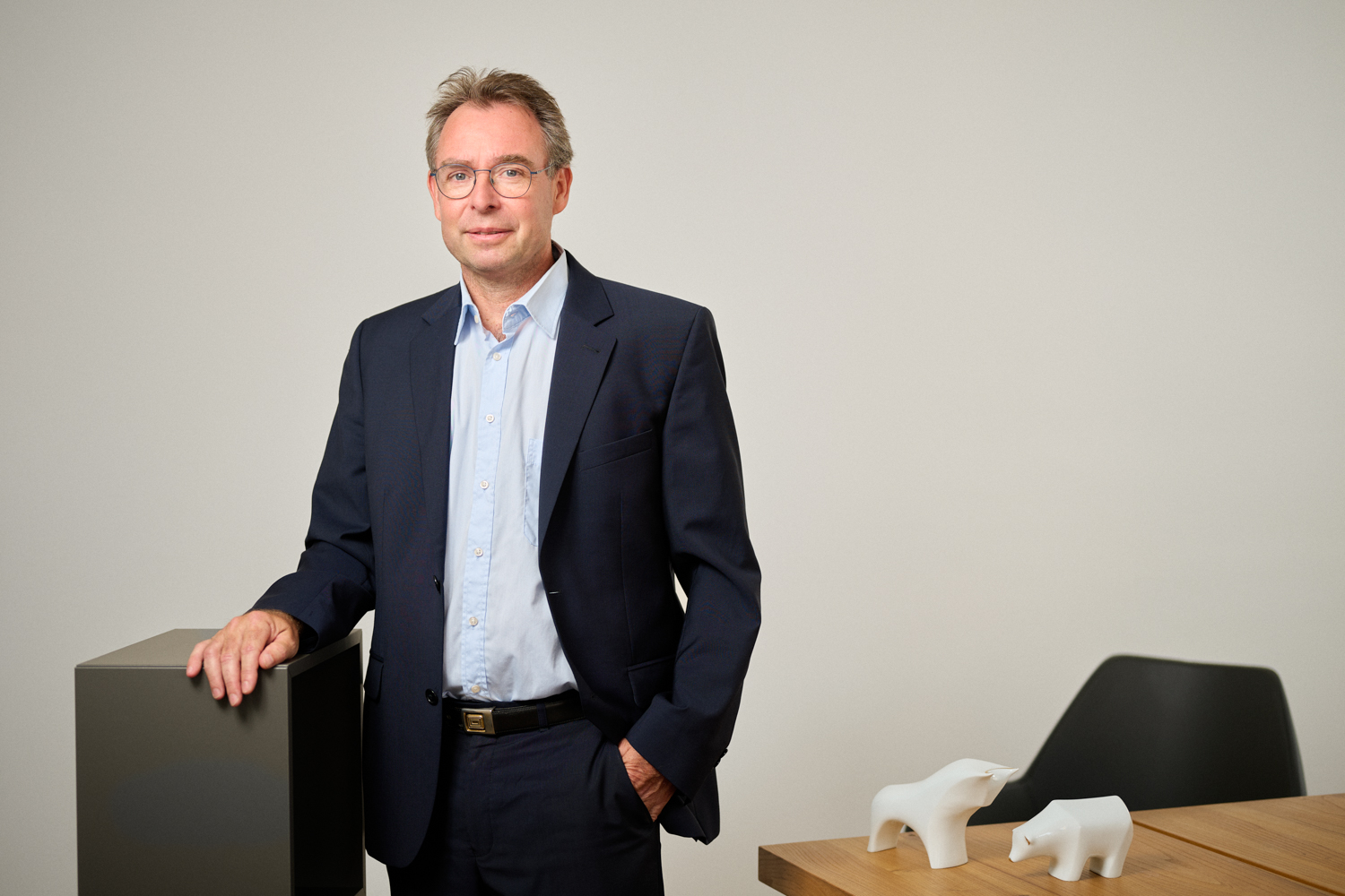 Portrait von Ralph Hofmann: Mann mittleren Alters mit kurzen braun-grauen Haaren und Brille, gekleidet in einen dunkelblauen Anzug mit hellblauem Hemd, lehnt mit einer Hand an einem dunklen Möbelstück in einem modernen Büro. Auf dem Tisch im Hintergrund stehen weiße Stier- und Bärfiguren.
