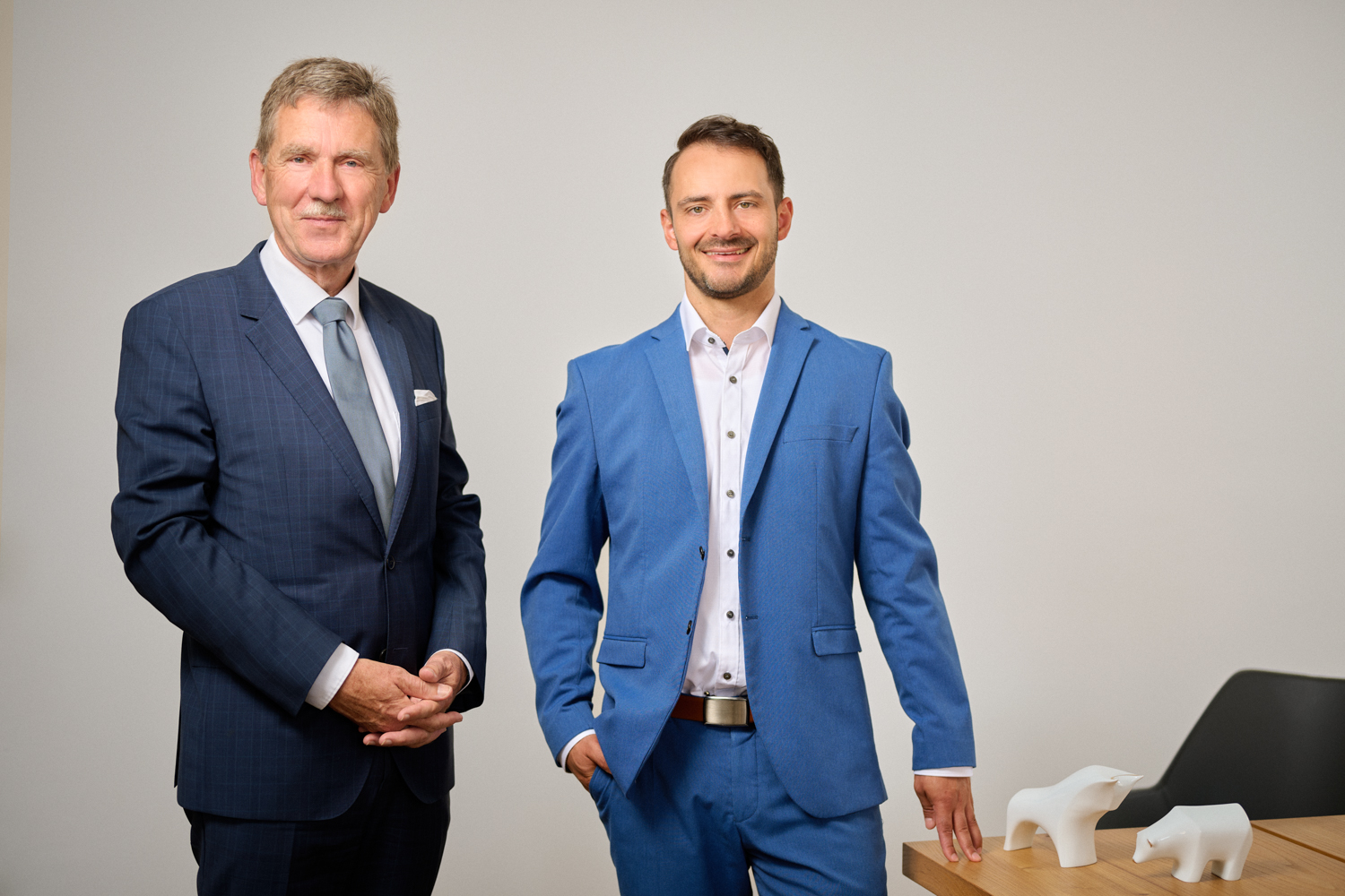 Portrait der beiden Vorstände der Fortezza Finanz AG. Links steht Wolfgang Dirsch, ein älterer Herr mit hellem Haar und dunkelblauem Anzug mit Krawatte. Rechts steht Alexander Stengel, ein junger Mann mit braunem, kurzem Haar und Dreitagebart, gekleidet in einem hellblauen Anzug. Beide lächeln freundlich in die Kamera.