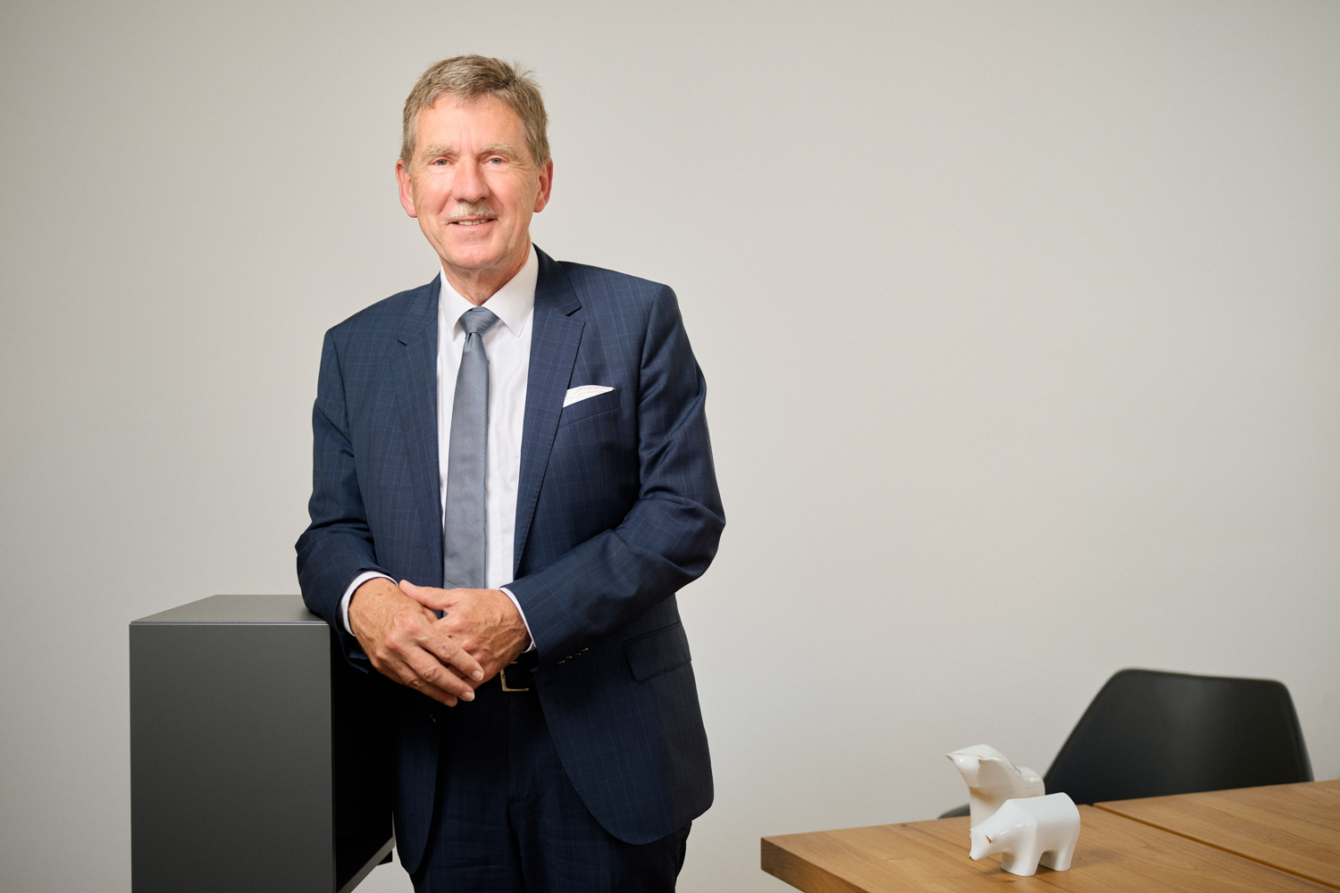 Portrait von Wolfgang Dirsch - Vorstand der Fortezza Finanz AG in Nürnberg: ein älterer Mann mit hellem Haar in dunkelblauem Anzug, weißem Hemd und Krawatte steht lächelnd in einem modernen Büro. Eine Hand ruht auf einem dunklen Möbelstück, im Hintergrund ein Tisch mit weißen Stier- und Bärfiguren.